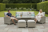 A man and a woman sit on a gray wicker outdoor patio conversation set with light gray cushions in a landscaped garden. The set includes a sofa, two armchairs, a rectangular coffee table, and two ottomans, arranged on a stone patio.