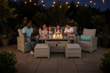 A family of four sits around a glass-top outdoor fire table with a wicker patio set on a stone patio at night, with warm string lights and plants in the background.