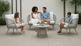 A family of four sits on a modern outdoor patio with four gray wicker armchairs with white cushions and a matching round coffee table. The background is a clean white wall with vertical dark gray accents and green plants.