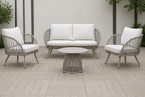 A 4-piece outdoor patio conversation set featuring a gray woven rope loveseat, two matching armchairs with white cushions, and a round coffee table, arranged on a light gray tiled patio.