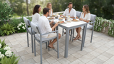 Group of people sitting around a dining table on a patio with greenery.