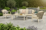 Outdoor patio set with beige furniture on a stone patio, surrounded by plants and flowers.