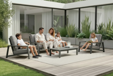Family sitting on outdoor furniture in a modern patio setting with plants and a building in the background.