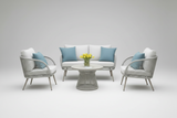 A modern outdoor seating set featuring a white woven sofa, two matching armchairs with light blue pillows, and a round woven coffee table with a vase of yellow flowers, set against a seamless light gray background.