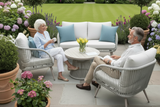 An outdoor garden patio scene with two people seated in woven armchairs facing a white sofa with blue pillows, arranged around a small round coffee table. The setting includes flower beds, potted plants, a manicured lawn, and a stone patio.